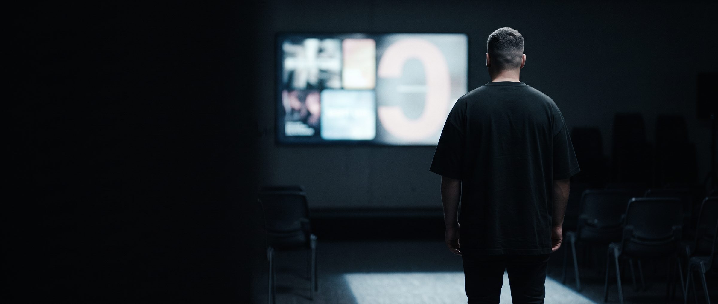 A man seen from behind in a dim room of empty chairs, looking at a large screen showing a grid of images