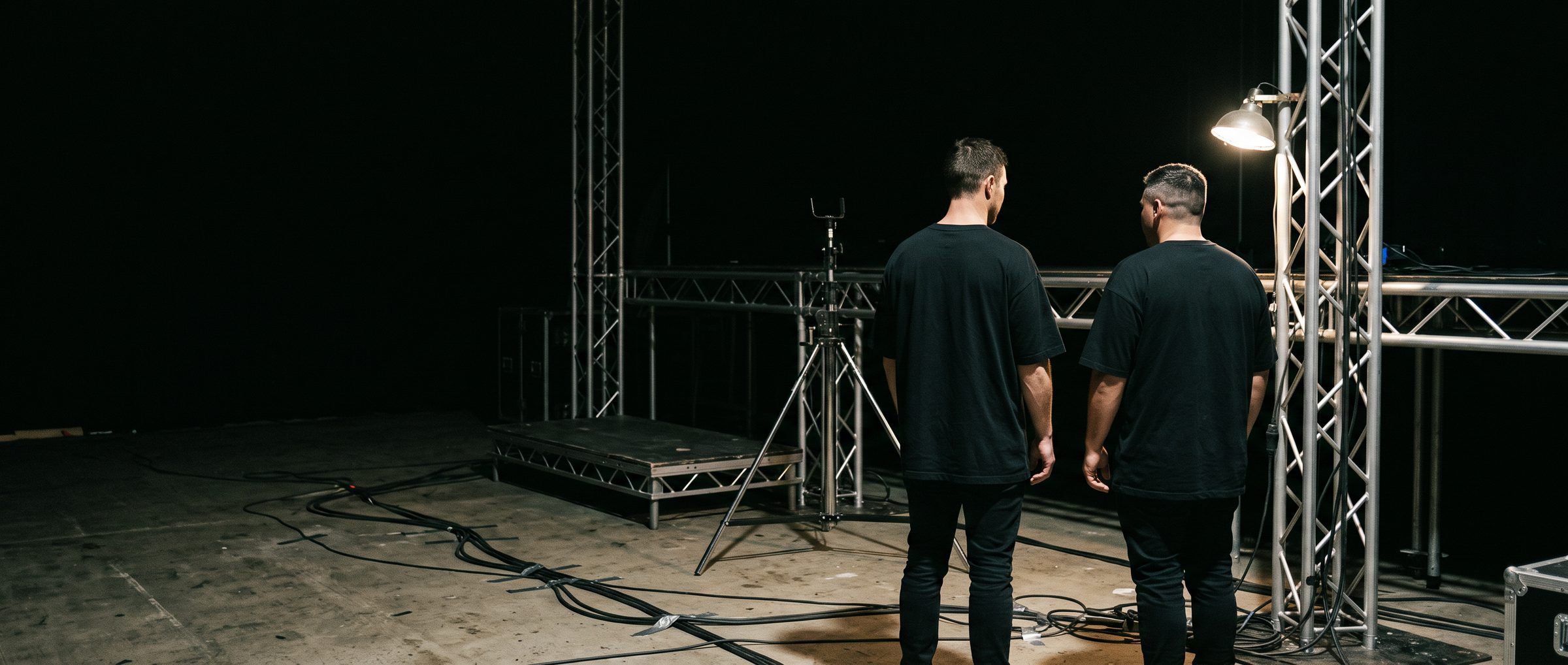 Two men seen from behind standing on a bare stage deck among aluminium truss, cables and a single work lamp