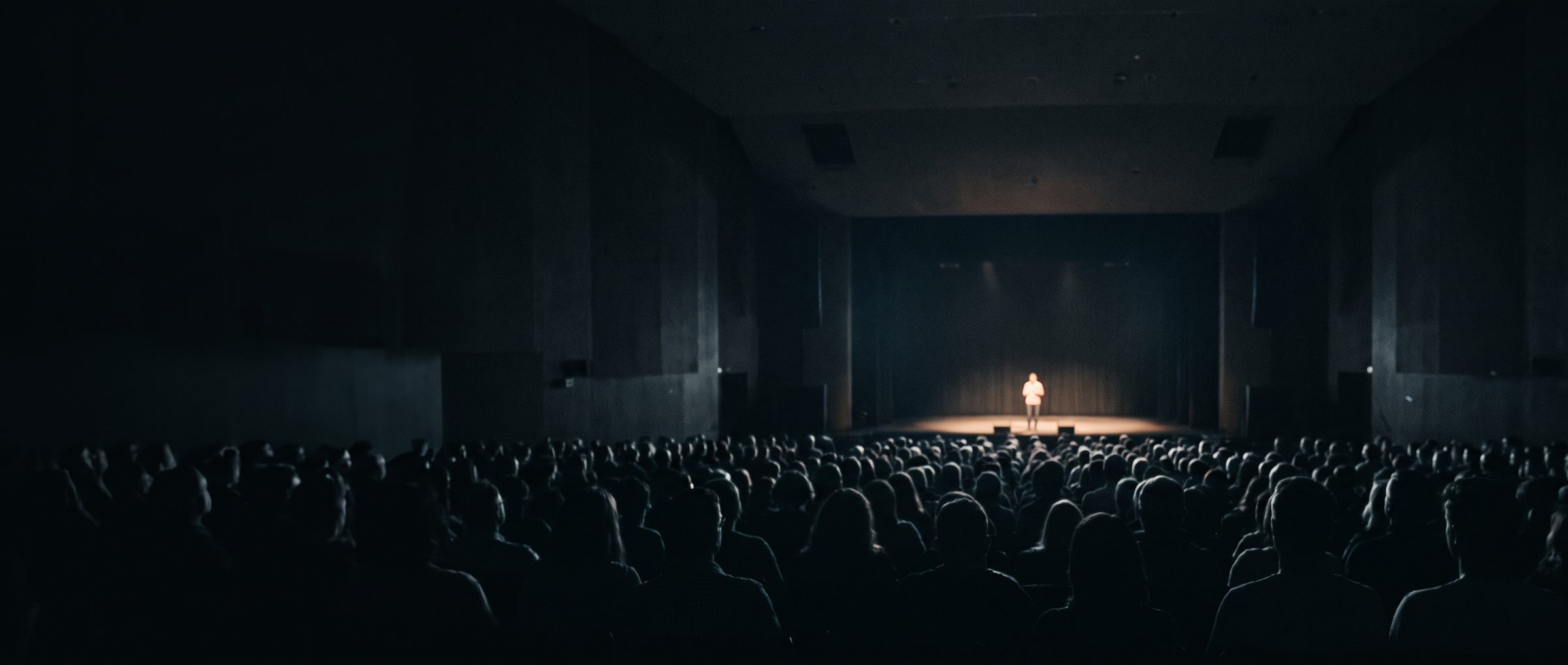 A packed, darkened theatre seen from the back rows, one speaker alone under a spotlight on a wide stage