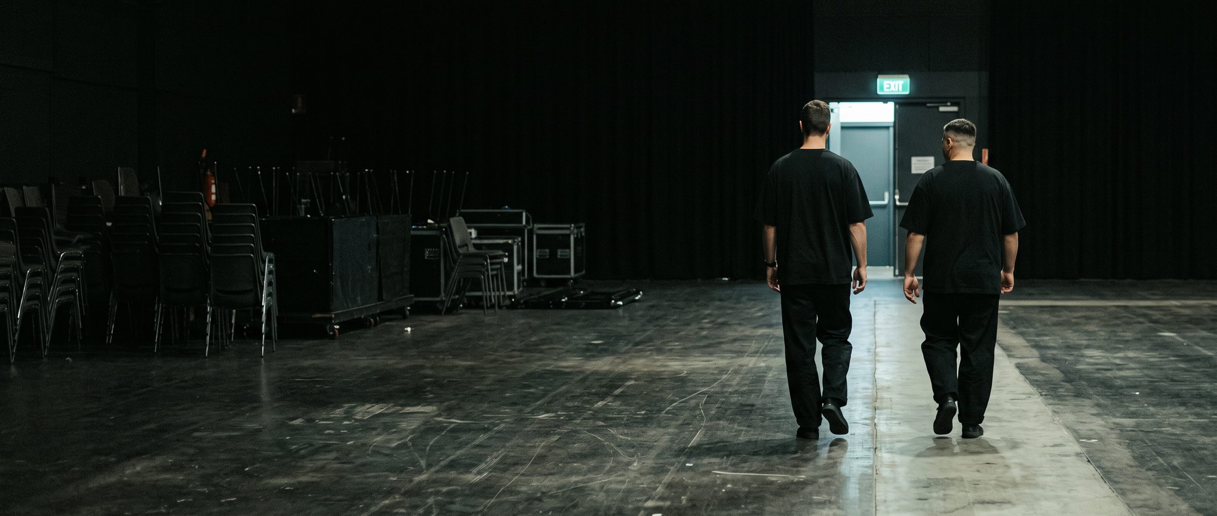Two men in black walking away across an empty venue floor towards a lit exit door, stacked chairs and road cases to one side
